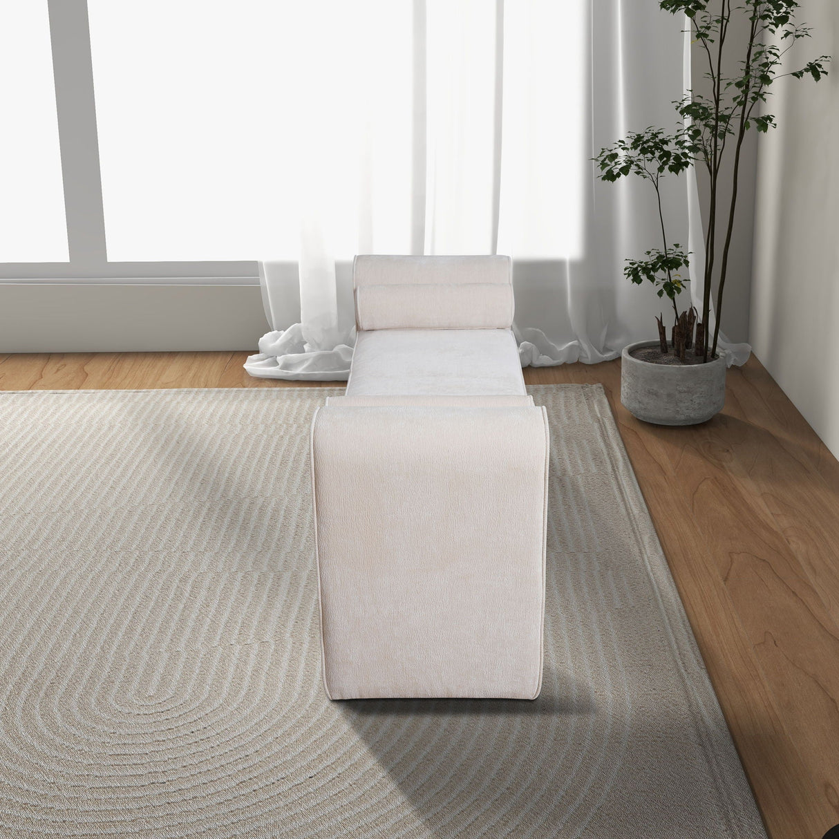 A minimalist room showcases the Regent Bench in cream, accented with a cylindrical pillow. A textured rug adorns the wooden floor, while a potted plant adds greenery. Natural light filters through sheer white curtains on the large window.