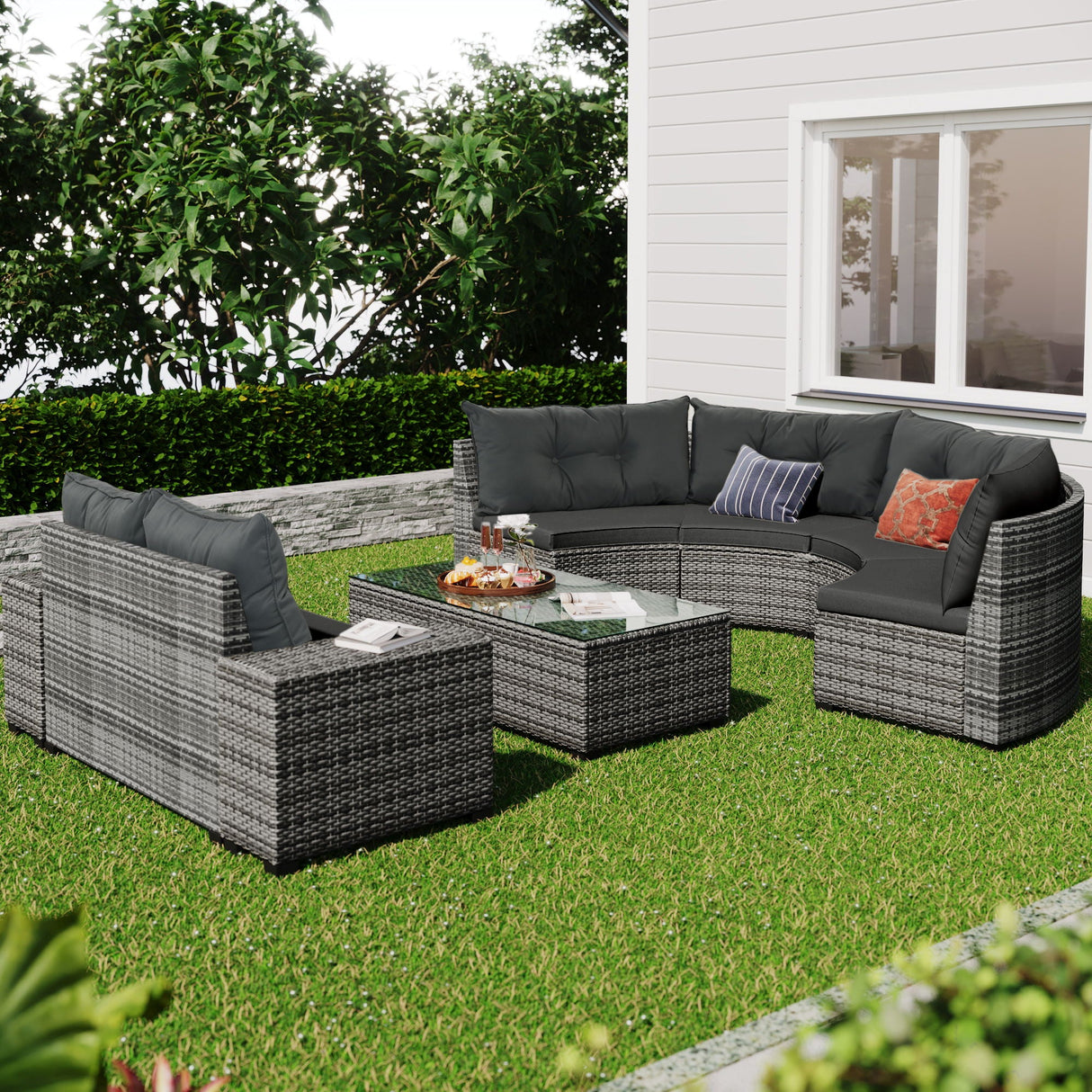 The outdoor patio features an 8 Pieces Outdoor Wicker Round Sofa Set, with a curved sofa and two chairs with dark cushions. A glass-top coffee table completes the set, all resting on grass. The area is framed by green hedges against a white house wall.