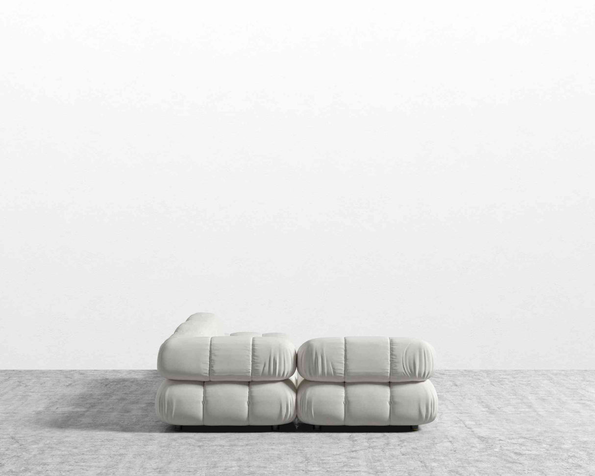 The Belia Modular Sectional, featuring tufted cushions in a light, soft fabric, is placed on a gray carpet against a white wall. The view from the back highlights its simple, modern design.