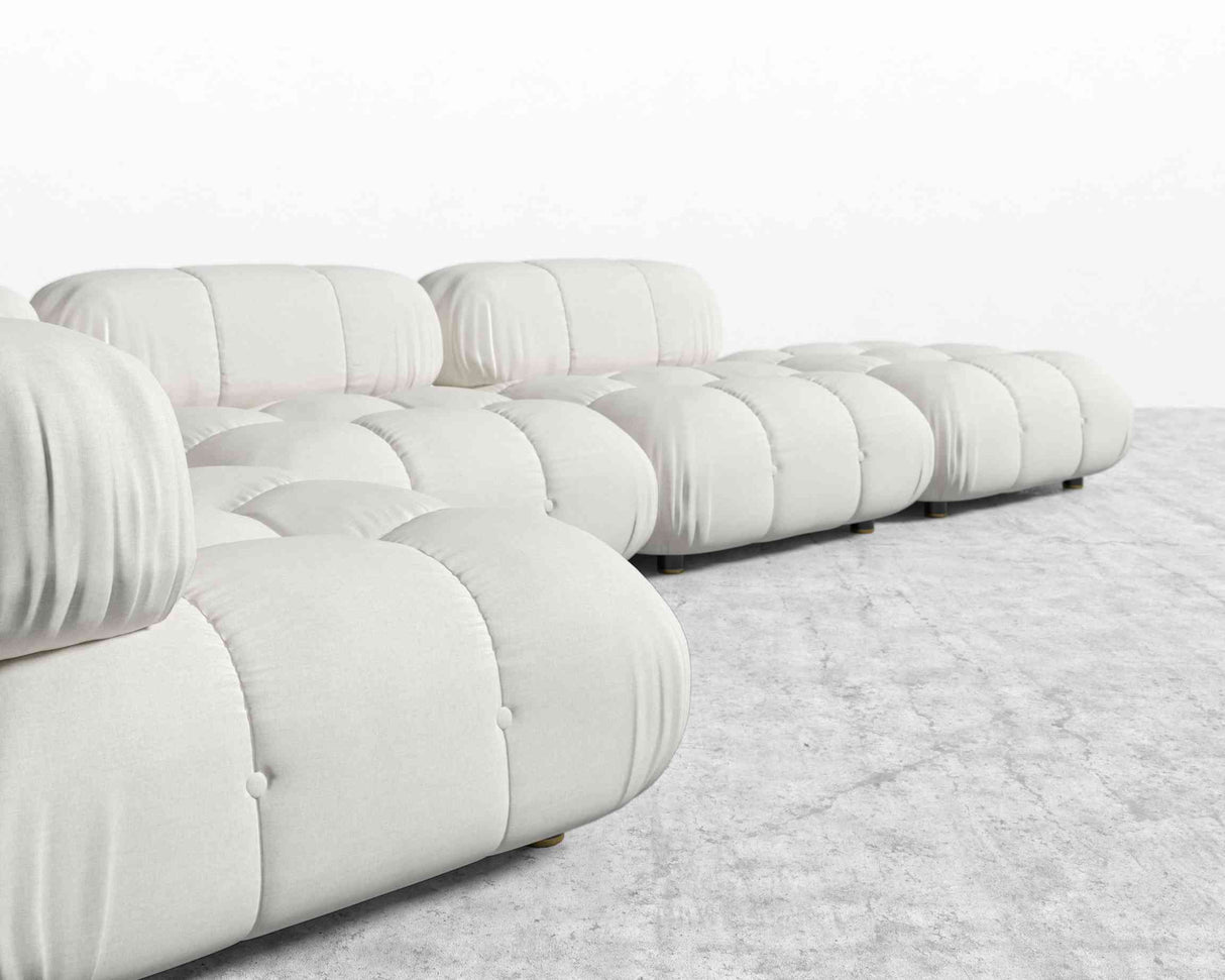 The Belia Modular Sectional in light gray upholstery is arranged on a matching light gray carpet. It features soft, rounded cushions with a tufted design, offering a modern and cozy look against the backdrop of a plain white wall.