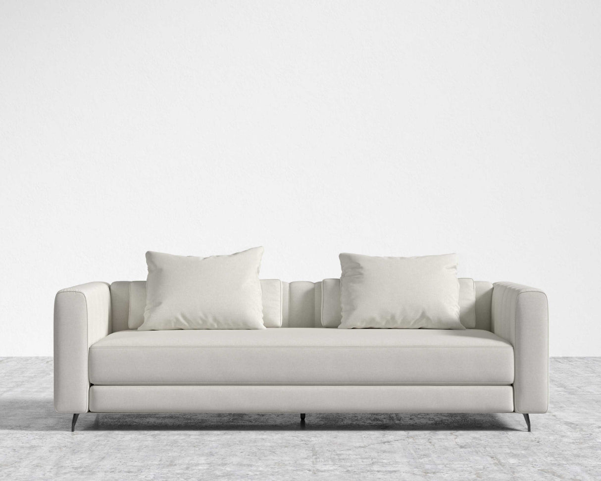 The Berlin Sleeper Sofa, featuring a modern and sleek minimalist design with smooth lines and subtle tufting on the arms, is placed against a plain white wall on a textured gray carpet. This light gray sofa comes with two matching square cushions.