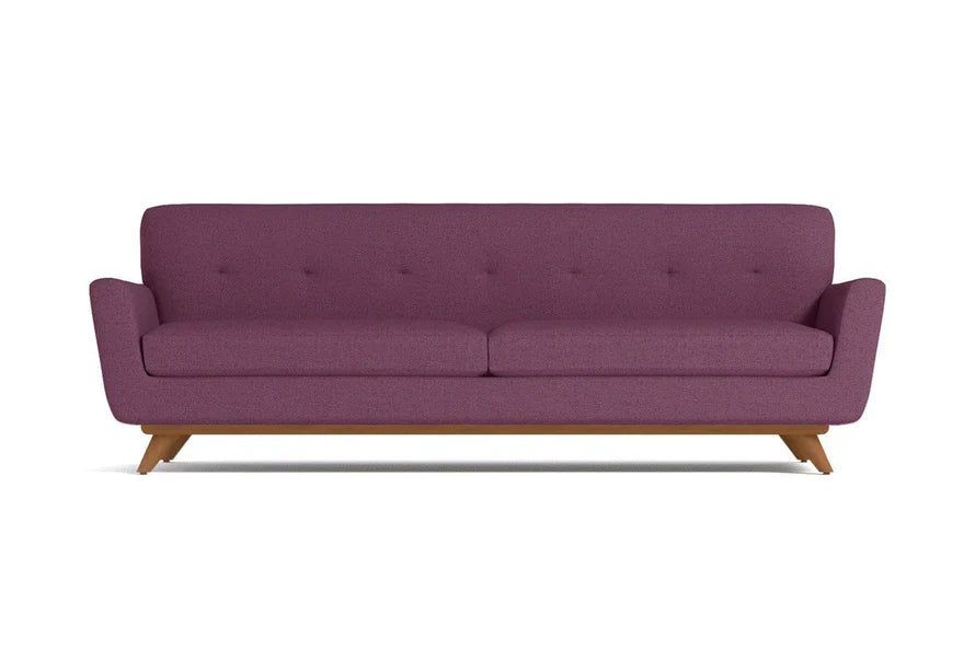 The Carson Sofa is a mid-century modern piece with purple upholstered fabric, featuring a tufted backrest and wooden legs. It boasts a minimalist design and comfortably seats three people.
