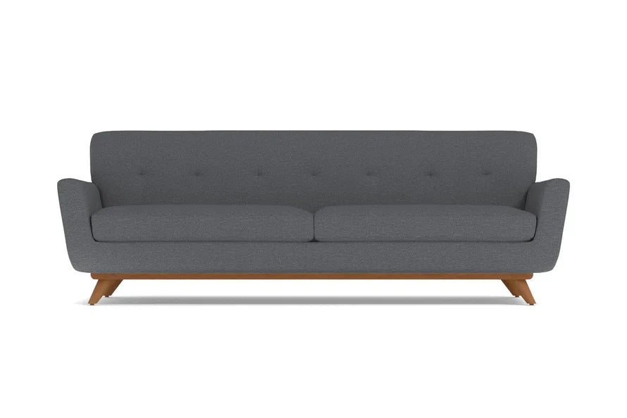 The Carson Sofa offers a modern aesthetic with its dark grey hue and minimalist design. It features a cushioned back adorned with subtle button detailing, sleek armrests, and is supported by mid-century styled wooden legs.