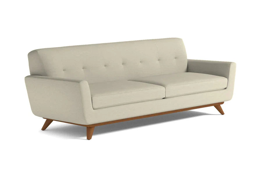 The Carson Sofa is a modern piece with tufted back cushions and wooden legs, showcased against a white background. It features a cream color, minimalist design with two seat cushions and slightly angled armrests.