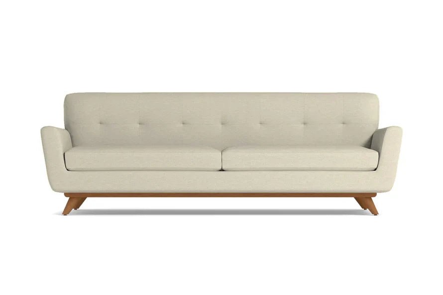 The Carson Sofa is a mid-century modern design featuring a beige color with a tufted backrest and two seat cushions. It is supported by a wooden base and angled legs, all presented against a plain white background.