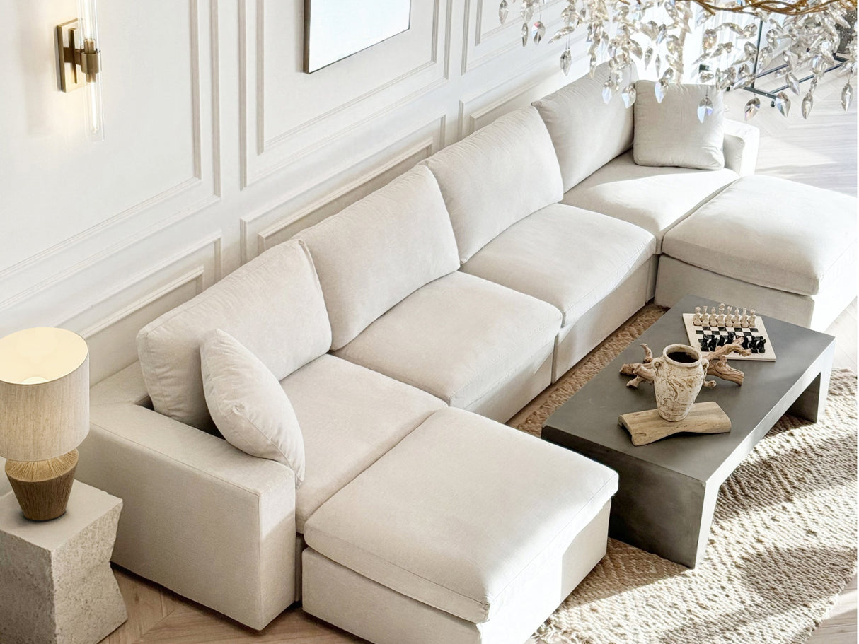 A spacious, beige 4-Seat Modular Chaise Sectional graces a well-lit living room with textured walls. A modern coffee table showcases decorative items, including a vase and chess set. Natural light streams in, highlighting the cozy, elegant ambiance.