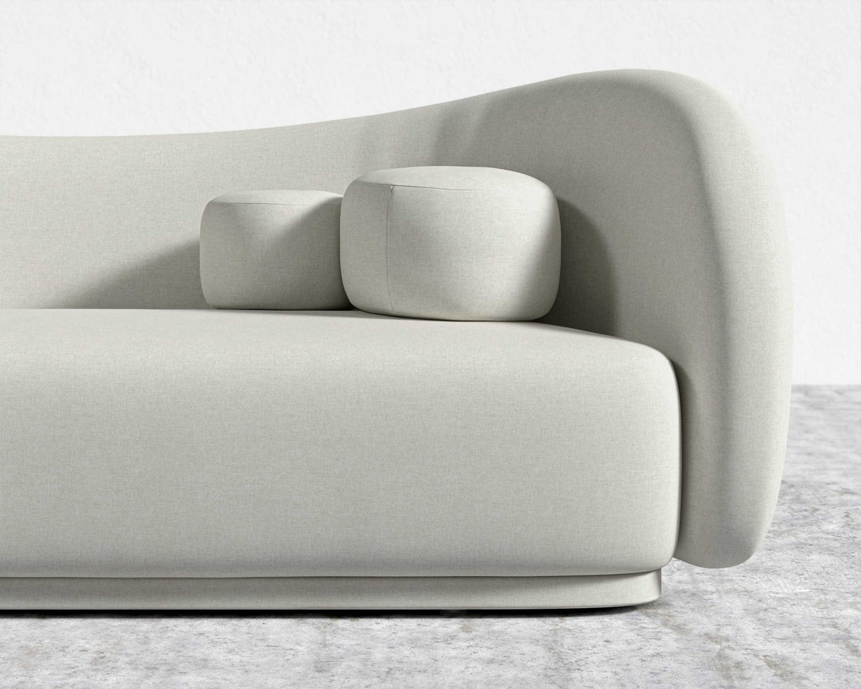 The Diane Sofa, a stylish and minimalist piece in beige, boasts a smooth curved design and is situated on a light gray textured floor. It includes two matching square cushions that enhance its modern aesthetic, set against a plain white wall backdrop.