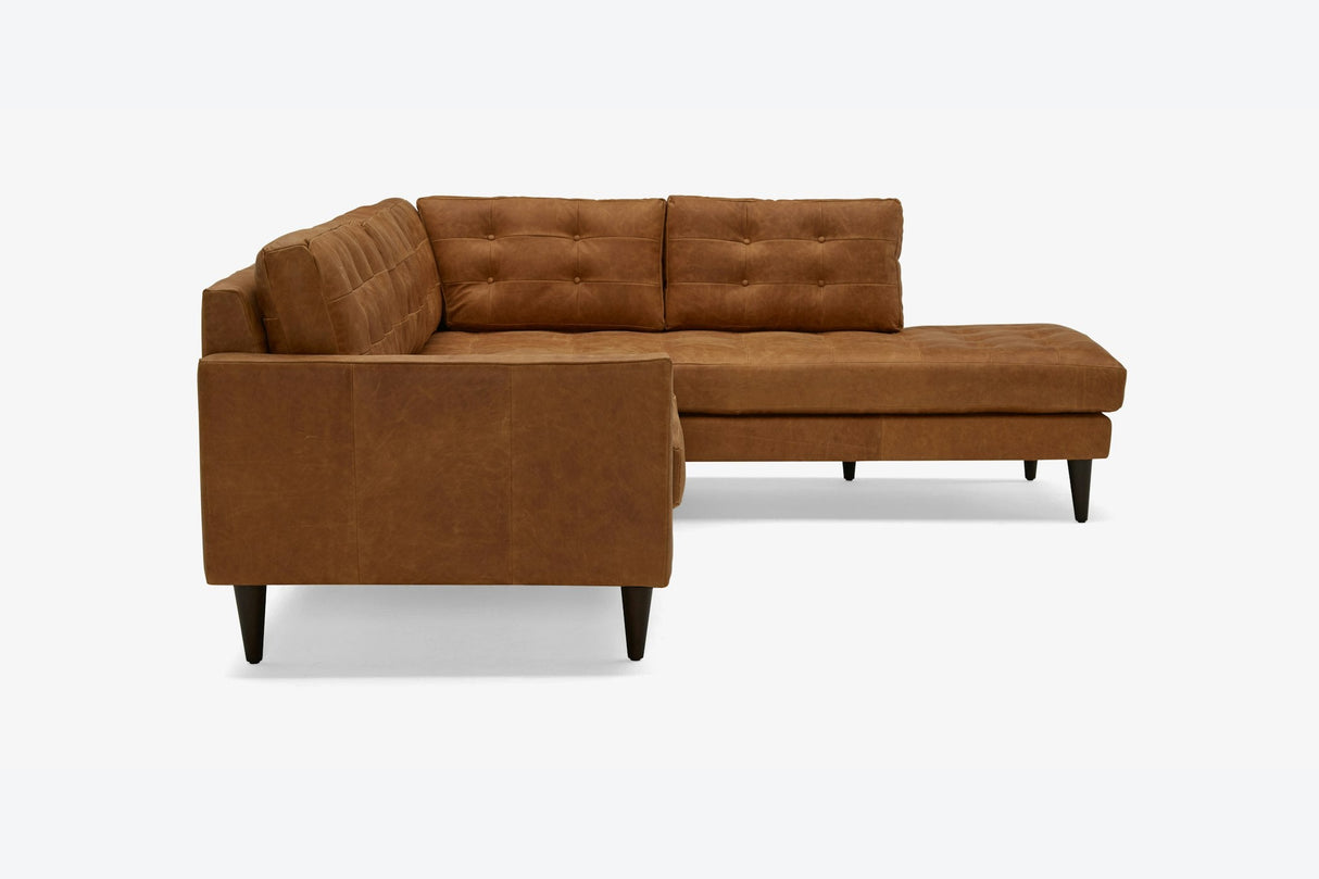 The Eliot Leather Sectional with Bumper, featuring a brown hue and L-shaped design, showcases tufted cushions and black legs against a light backdrop. This sofa offers a modern aesthetic with generous seating space.