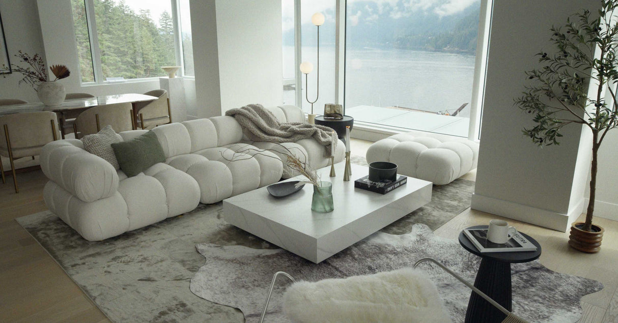 A modern living room showcasing the Belia Modular Sectional in white, complemented by matching ottomans and a sleek white coffee table decorated with accents. Large windows offer stunning views of a lake and forest, while a floor lamp and a plant enhance the cozy ambiance.