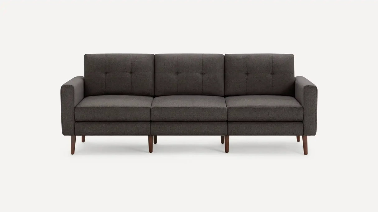 The Nomad Fabric Sofa, a modern three-seater in dark gray with square cushions and wooden legs, is showcased against a plain white background. It boasts clean lines and a minimalist design.