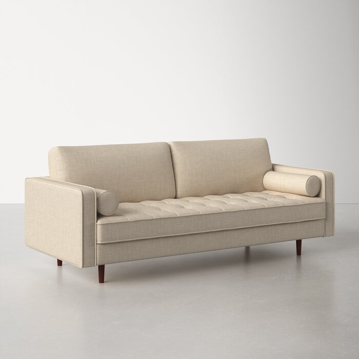 The Geo 84'' Upholstered Sofa in beige, featuring tufted cushions, cylindrical arm pillows, and wooden legs, is showcased on a light gray floor with a white background.