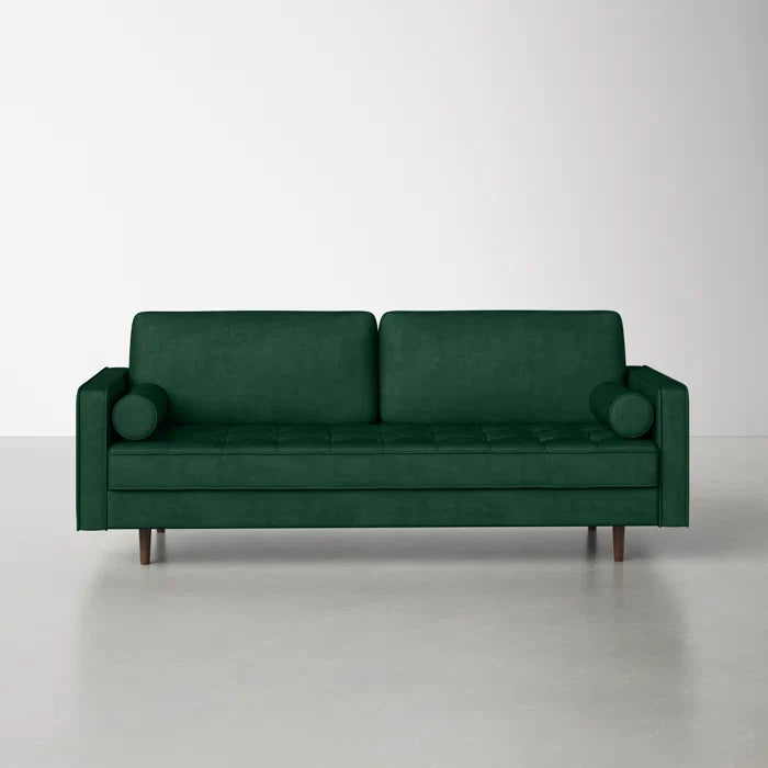 The Geo 84'' Upholstered Sofa, featuring a modern design with green velvet upholstery, includes two seat cushions and two cylindrical bolster pillows. It stands in an empty room with a light-colored floor and wall, supported by four wooden legs that enhance its minimalist aesthetic.