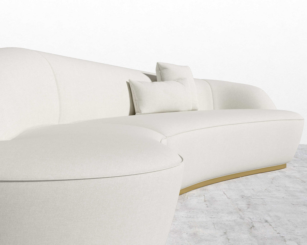A contemporary Reya Curved Sectional in white, complemented by two matching square cushions and placed on a light gray textured rug with a white backdrop. The sofa showcases a minimalist design with a smooth, elegant finish.