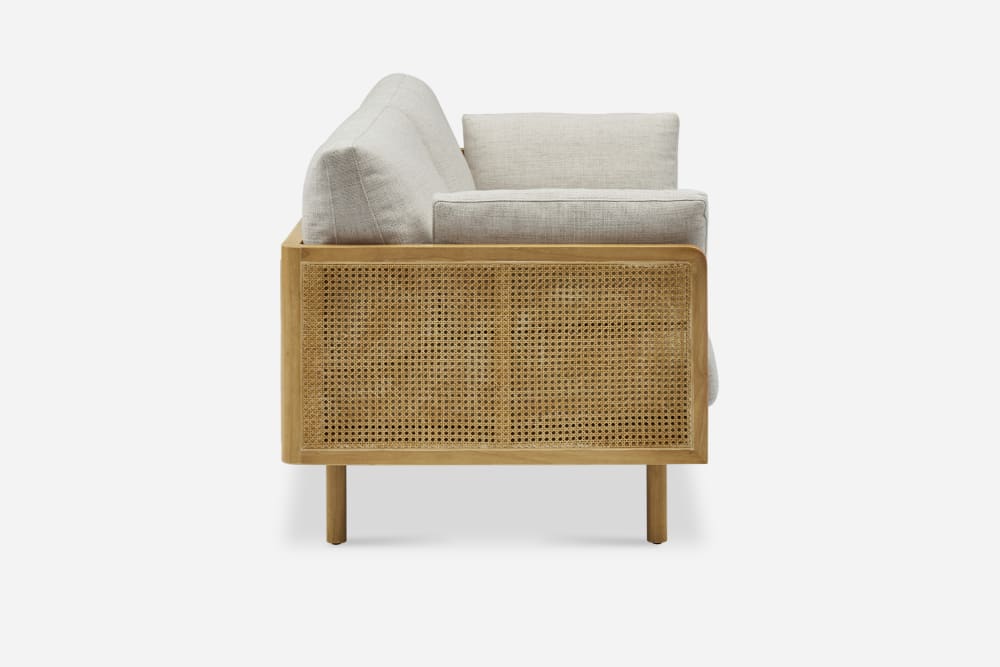 Side view of the Lucia Cane Sofa, White Wash, featuring light beige cushions and a wooden frame with a woven rattan design on the side panel, set against a plain white background.