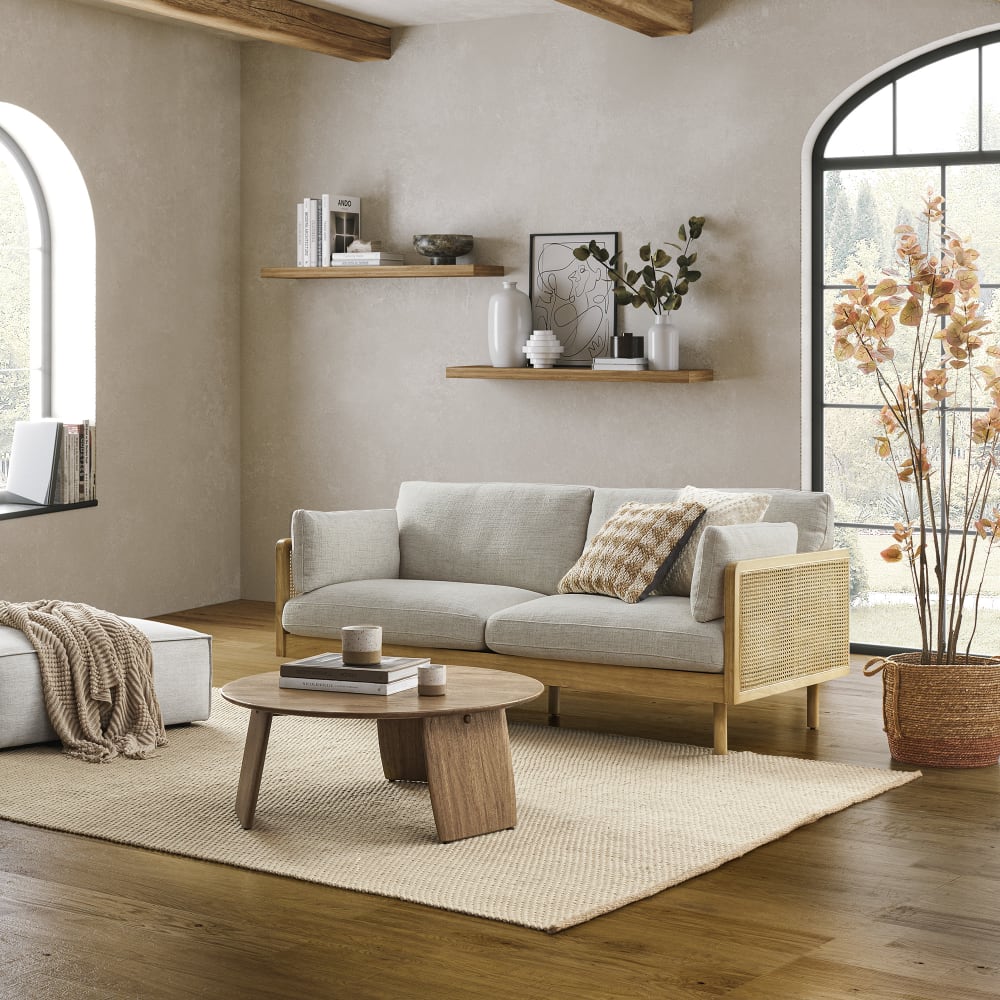 A cozy living room features a Lucia Cane Sofa in White Wash, accompanied by a wooden coffee table and a decorative pouf. Shelves filled with decor items adorn the wall, while a large window allows natural light to pour in. A tall plant with autumn-colored leaves enhances the warmth of the room.