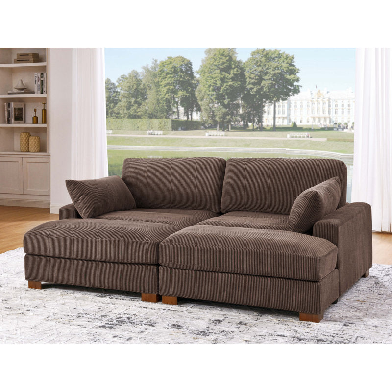The Etta Avenue Mordecai 90'' Square Arm Sofa, featuring plush cushions, is placed in a living room with a large window that offers a scenic view of a park with trees and a distant white building. A bookshelf and patterned rug further enhance the cozy atmosphere.