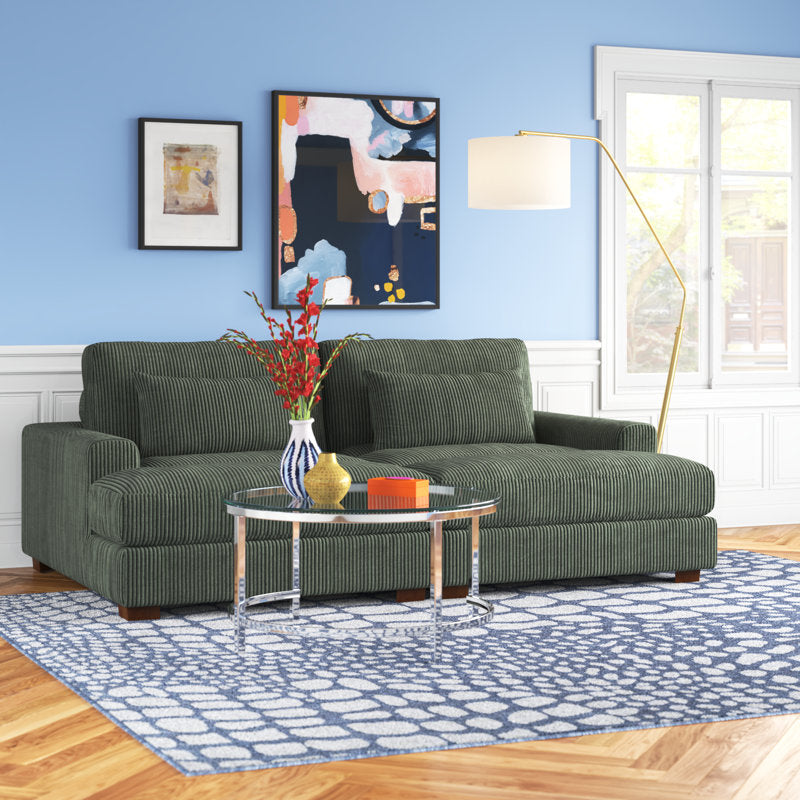 A cozy living room features the Etta Avenue Mordecai 90'' Square Arm Sofa in green corduroy, placed on a blue patterned rug. A round glass coffee table displays a vase of red flowers and books. Abstract art adorns the blue wall, while a tall floor lamp brings warmth to the space.