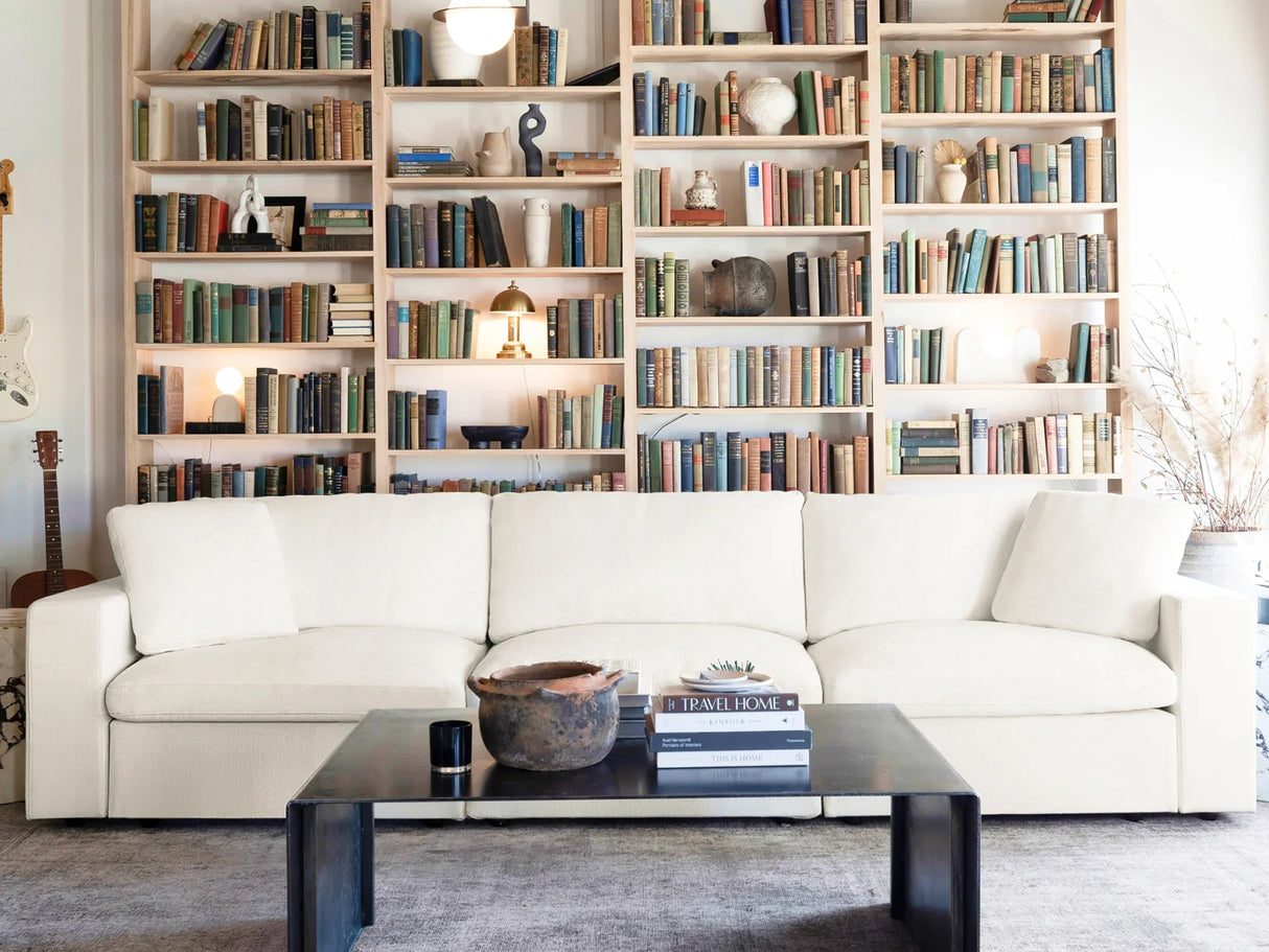 In a cozy living room, a large 4-seat modular chaise sectional with ottoman is situated in front of floor-to-ceiling bookshelves filled with assorted books. A black coffee table holds a stack of books and a rustic bowl, while soft lighting and a guitar accentuate the inviting space.