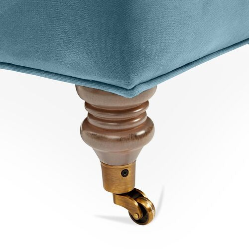 Close-up of a Brooke 6-Seat Sofa leg featuring a wooden turned design with a brass caster wheel. The light blue fabric upholstery above exudes a classic and elegant style.