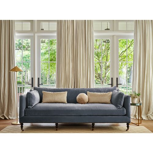 In a cozy living room, the Margot Velvet Sofa in gray is adorned with beige cushions and set against large windows framed by cream curtains. A gold floor lamp and a round side table complete the elegant and serene setting.