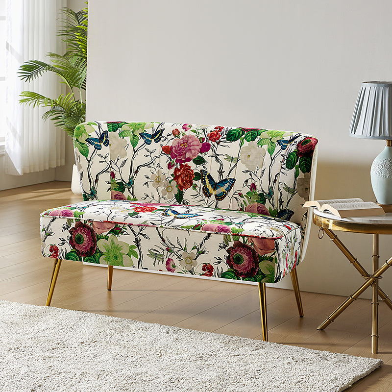 The Coraline Loveseat, featuring a vibrant floral and butterfly design, rests elegantly on a light wooden floor. Beside it stands a small round table adorned with a blue lamp and a book. Sunlight pours through sheer curtains, casting light onto the scene as a potted plant adds greenery to the background.