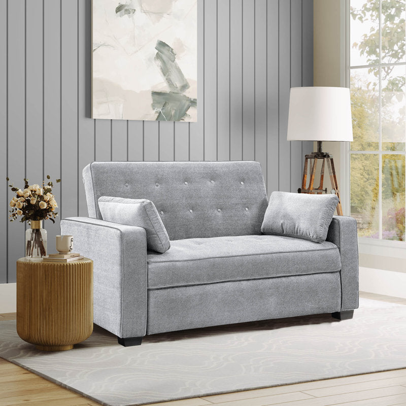 A cozy living room features a Serta Monroe Full Size Convertible Sofa in light gray with button tufting and matching cushions. A wooden side table holds a vase of flowers, accompanied by a modern floor lamp near a large window showcasing a view of greenery. Wall art decorates the gray-paneled wall.