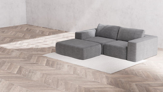 A minimalist room showcasing a light gray Modular 2-Seater Chaise Sectional in Boucle Aspen, complemented by an ottoman, is set against a cream-colored rug. The area boasts herringbone-patterned wooden flooring and light gray walls, establishing a serene and stylish ambiance.