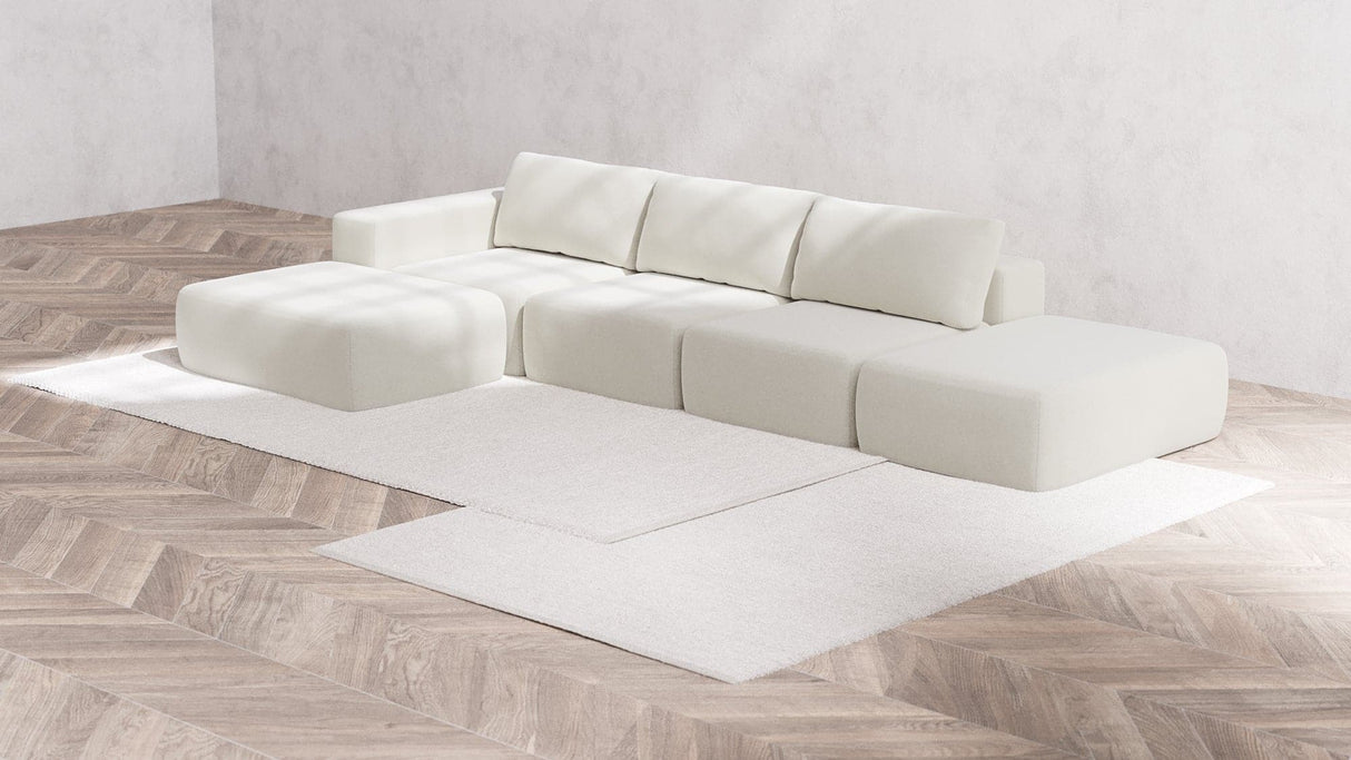 The Modular 3-Seater Open-End Chaise Sectional in Boucle, Aspen, with its modern and modular design featuring clean lines, is arranged in an L-shape on a light wood herringbone floor. Two white area rugs are layered underneath, creating a minimalist and serene living space.