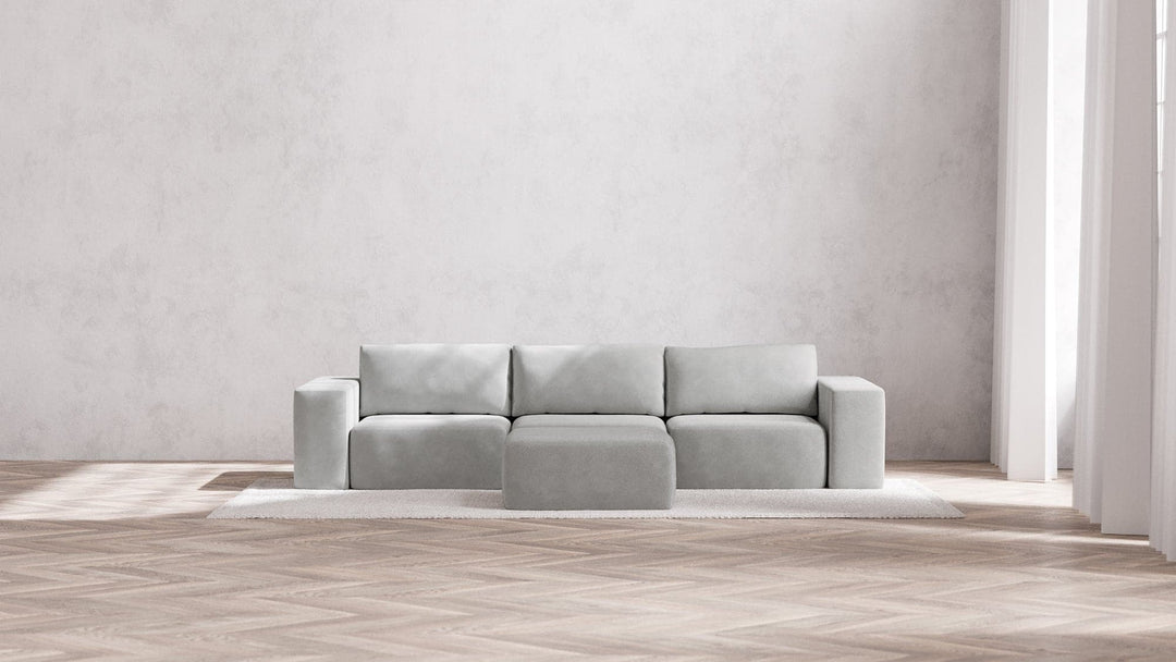 A minimalist, modern living room featuring the light gray Modular 3-Seater Sofa + Ottoman in Boucle Aspen atop a textured rug, set against a plain gray wall. Natural light pours in through tall windows, highlighting the herringbone-patterned wooden floor.