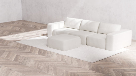 A minimalist living room showcases a modern Modular 3-Seater Sofa and Ottoman in Boucle Aspen, set on a textured rug. The room features light wood herringbone flooring and neutral walls, creating a clean, airy atmosphere.