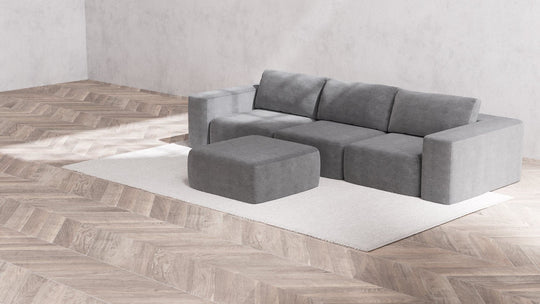 In the minimalist living room, a large Modular 3-Seater Sofa and Ottoman in Boucle Aspen sits atop a light beige rug. The herringbone-patterned wooden flooring complements the plain gray walls. The space is well-lit, resulting in a cozy and modern atmosphere.