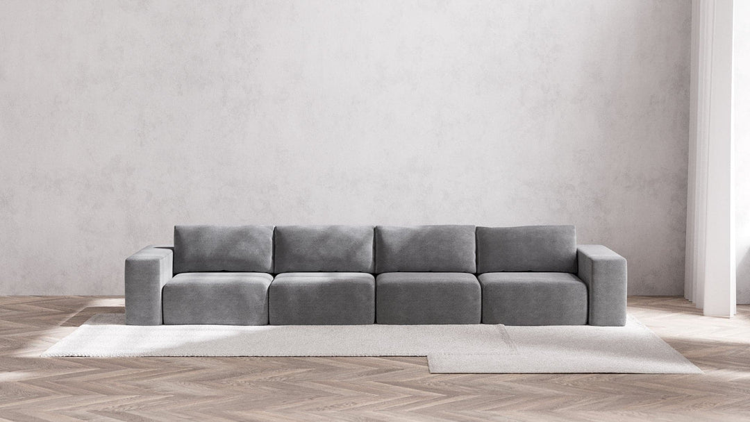 A modern, minimalist living room showcases the Modular 4-Seater Sofa in Boucle Aspen, positioned against a light gray wall. The sofa rests atop a large, light-colored area rug laid over a wooden herringbone floor, cultivating a serene and airy ambiance.