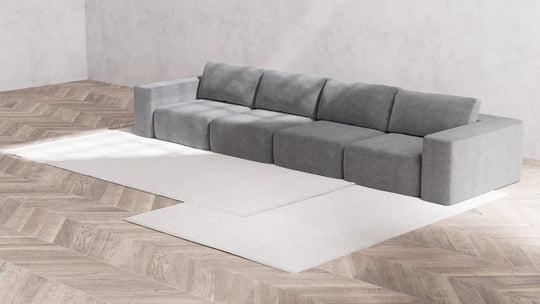 A modern, Modular 4-Seater Sofa in Boucle Aspen sits in a minimalist living room with wooden herringbone flooring. Two white, rectangular rugs are placed partially under the sofa. The walls are bare and white.