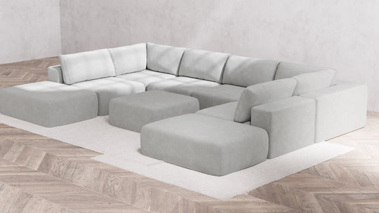 A contemporary, minimalist corner sofa styled as the Modular 8-Seater Open-Ends U Sectional + Ottoman in Boucle Aspen graces a white rectangular rug. It features a modular design accompanied by matching ottomans. The flooring showcases light herringbone wood, complemented by plain gray walls.