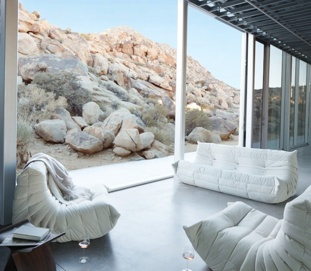 A modern living room featuring the Togo Sofa in Opera Leather and large glass windows offers a view of a rocky desert landscape. Sunlight brightens the space, accentuating the sleek white flooring and minimalist decor, with a couple of glasses resting on the floor.