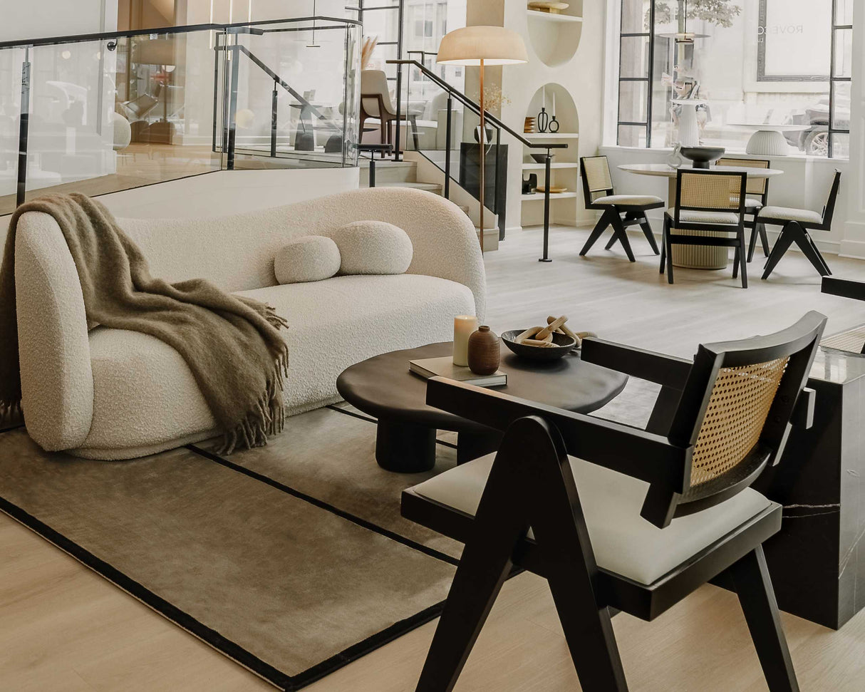 A sophisticated living room features a cream-colored Diane Sofa with a dark wood coffee table and modern black chairs. A cozy throw is draped over the sofa. Large windows illuminate the space, revealing a dining area in the background.