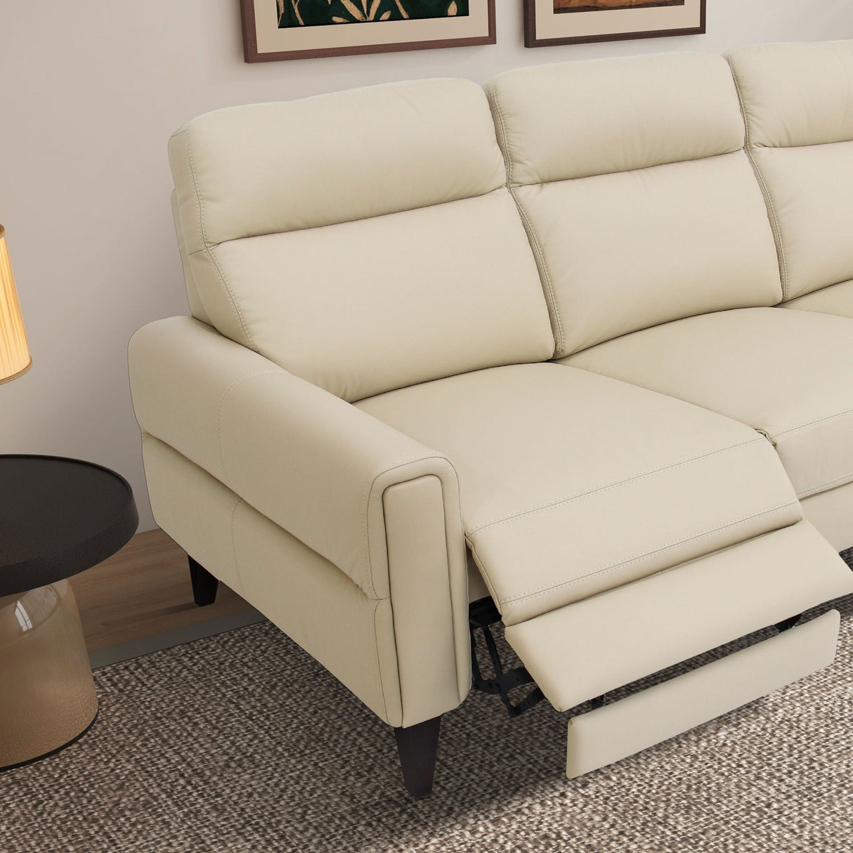 The Elliot Incliner Sofa in beige, with an extended footrest, rests on a textured rug. A black round side table and a warm-toned lamp accompany it, while partial wall art is visible above.