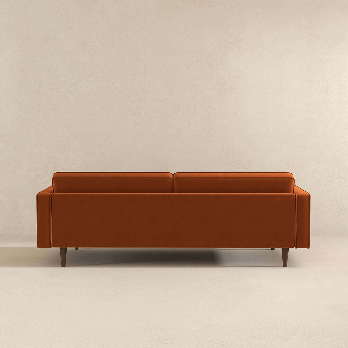 The Casey - Mid Century Modern Sofa, with its sleek rectangular shape and wooden legs, is a minimalist orange piece set against a plain light-colored wall, featuring clean lines and a backrest that complements its contemporary style.
