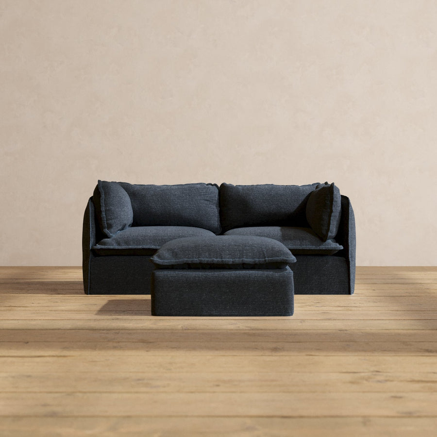 A Modular Performance 2-Seater in dark blue, complete with matching cushions, is positioned on a wooden floor against a beige wall. A small square ottoman in the same color is placed in front of the sofa.