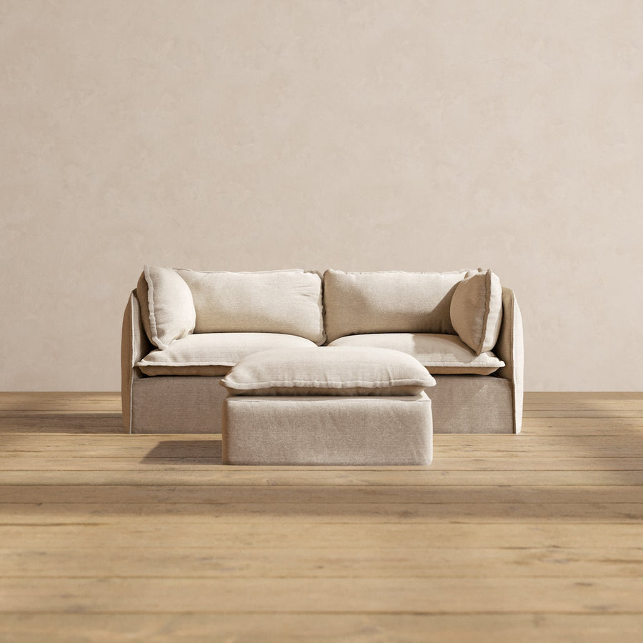 A Modular Performance 2-Seater in minimalist beige, complete with plush cushions, sits on a wooden floor. A matching ottoman is positioned in front of the seating arrangement. The background showcases a neutral cream-colored wall.