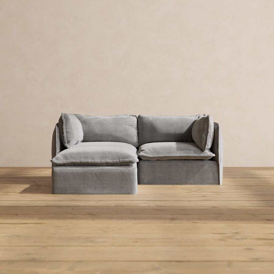 A Modular Performance 2-Seater Sectional in modern gray, featuring plush cushions, is set against a neutral beige wall. Positioned on a wooden floor, it creates a minimalist and cozy ambiance.