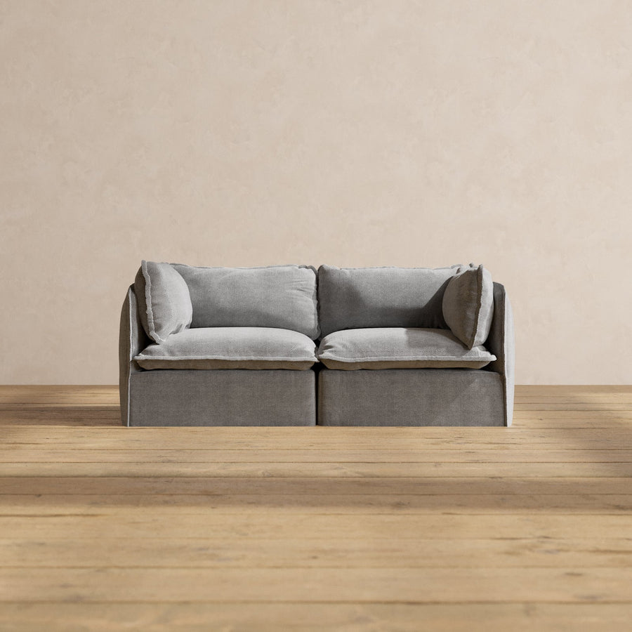 A Modular Performance 2-Seater with Sink-in Comfort Cushion in a modern gray design rests on a wooden floor. The plain beige wall in the background enhances the minimalist and cozy ambiance.