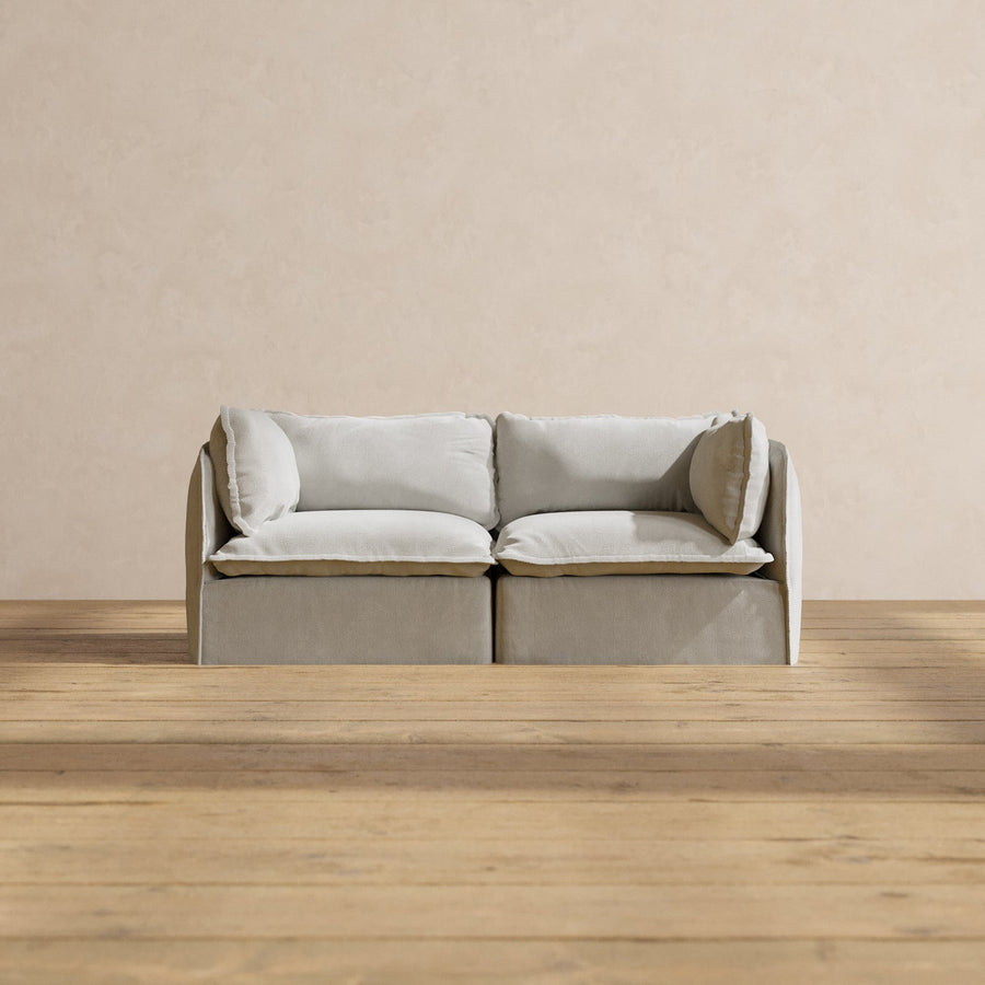 A minimalist room with a light gray Modular Performance 2-Seater featuring Sink-in Comfort Cushion on a wooden floor. The sofa boasts plush cushions, and the wall behind is plain and textured in a neutral tone.