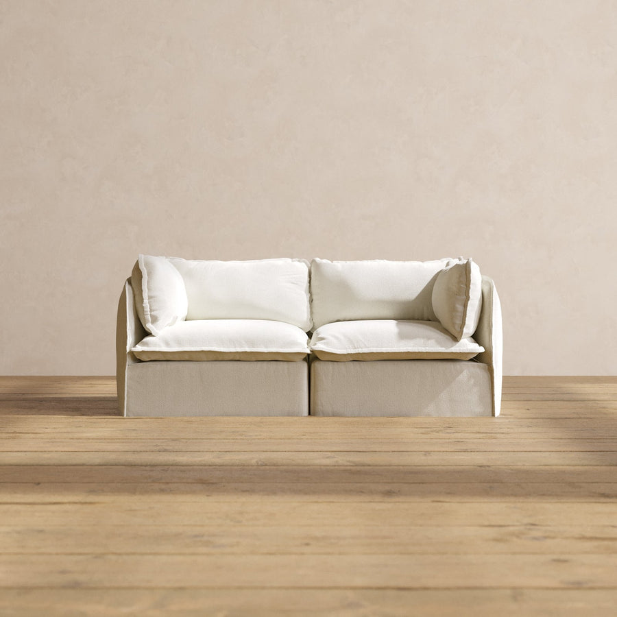 A minimalist, modern white Modular Performance 2-Seater with Sink-in Comfort Cushion sits on a wooden floor against a plain beige wall. The sofa features two large cushions and a sleek design, creating a serene and contemporary atmosphere.