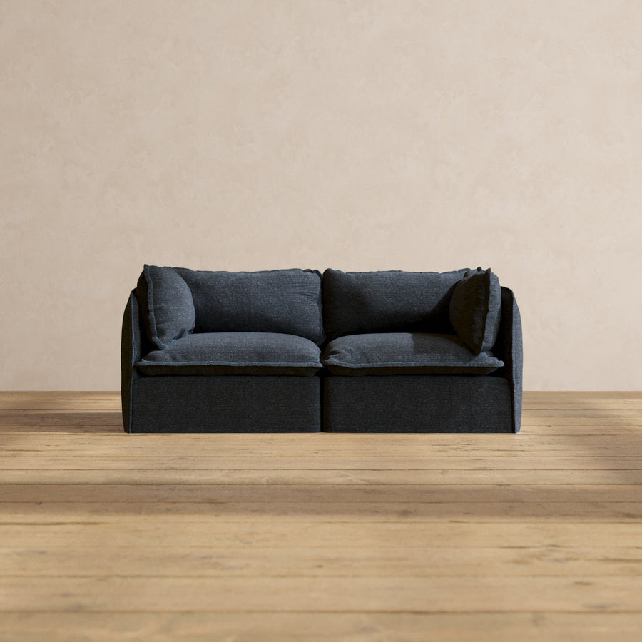 A Modular Performance 2-Seater with Sink-in Comfort Cushion, featuring a dark gray finish and two large cushions, is situated on a wooden floor against a light beige wall. The sofa showcases a minimalist and modern design.