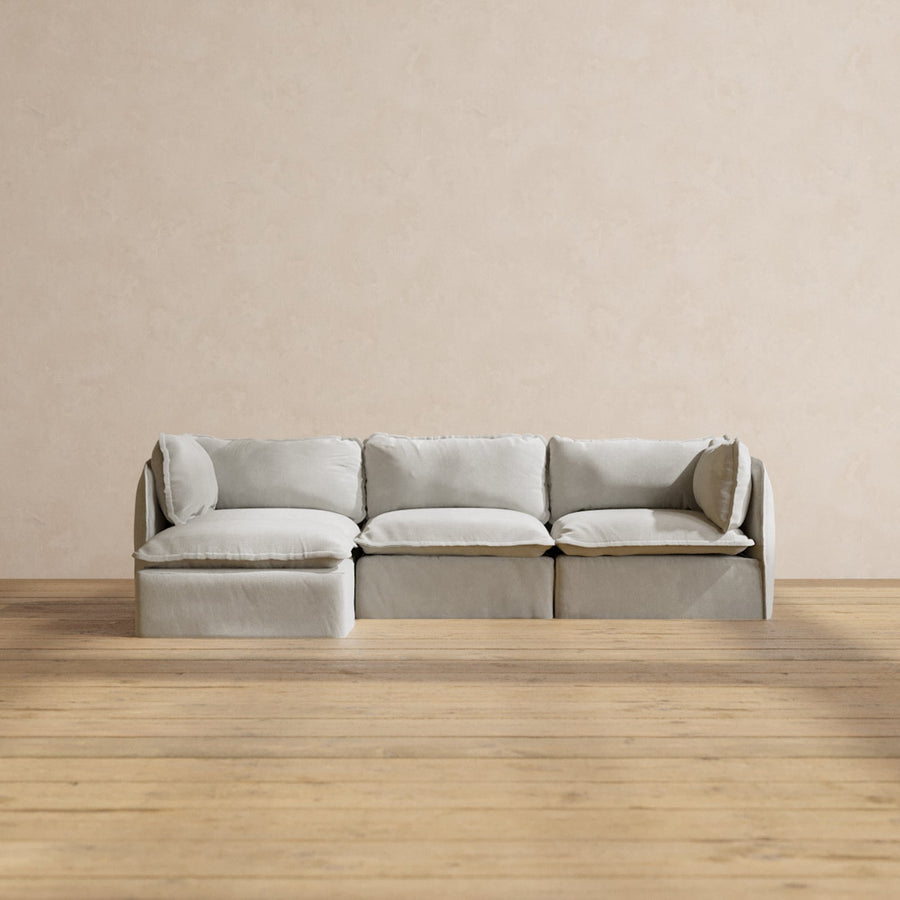 A minimalist Modular Performance 3-Seater Sectional in light gray, featuring a chaise lounge on the left side, is positioned against a neutral beige wall. The room's wooden floor adds a warm contrast to this simple, elegant setting.