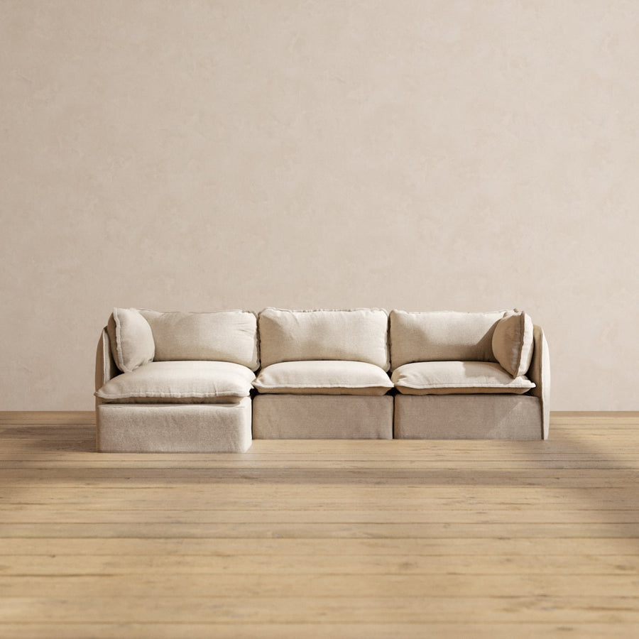 A Modular Performance 3-Seater Sectional in beige, complete with four cushions, is situated in a room featuring light wooden flooring and a subtly textured cream wall. The sofa showcases a modern, minimalist sectional design.