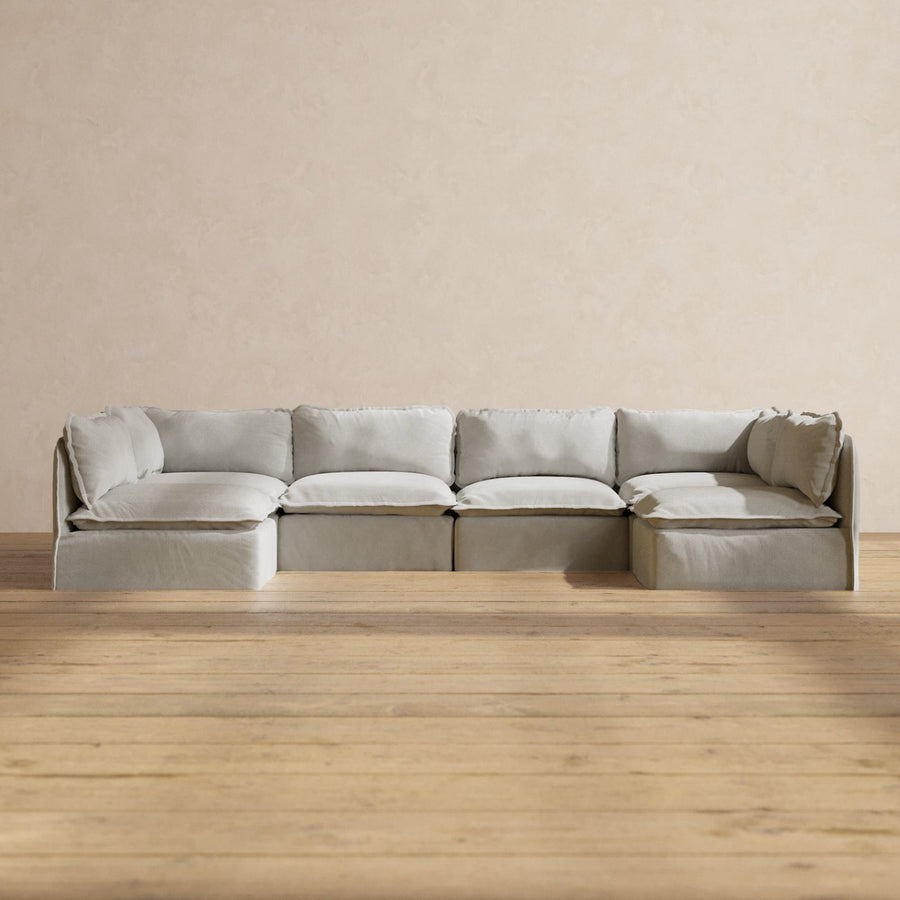 A Modular Performance 6-Seater Open-Ends U-Sectional in Plush Weave Ash is configured in an L-shape on a wooden floor, set against a light beige textured wall.