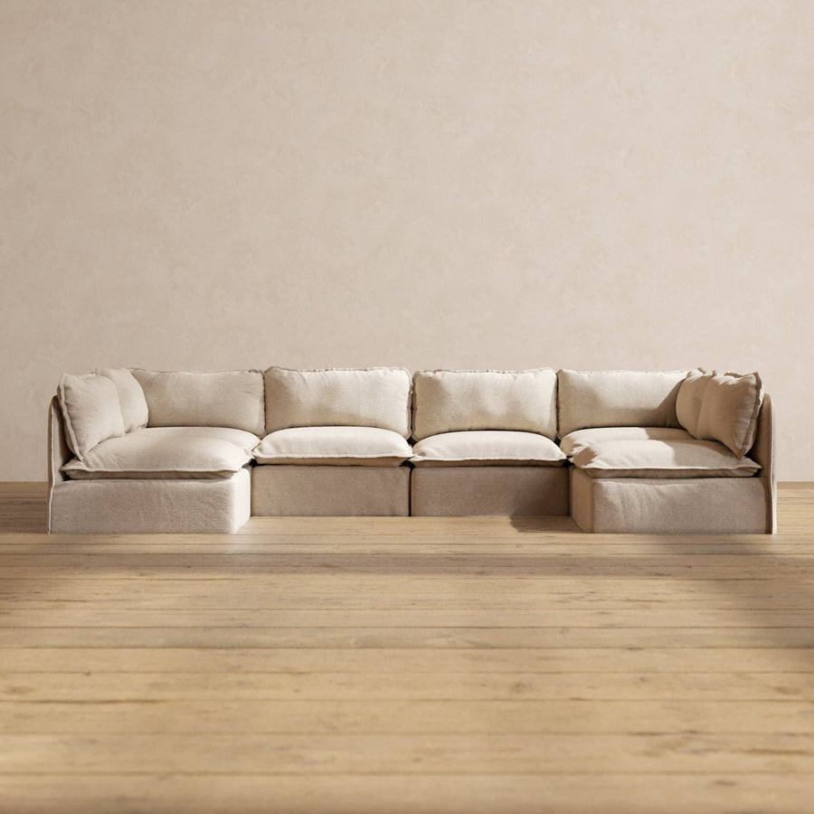 The Modular Performance 6-Seater Open-Ends U-Sectional in Plush Weave Ash features thick cushions and is set against a plain, light-colored wall. The room has light wood plank flooring that enhances the warm and minimalistic ambiance.