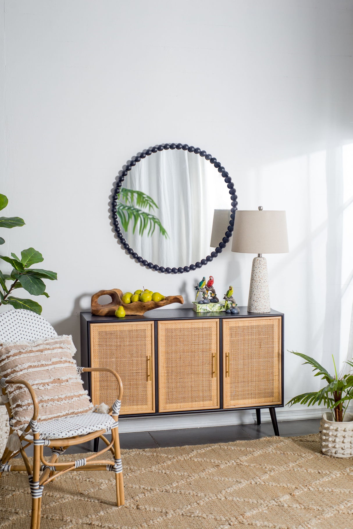 A room features a rattan chair with a cushion, a sideboard with woven doors adorned by the Circle Mirror With Metal Beaded Frame in black above it. A lamp, decorations, and lemons sit on the sideboard. A potted plant and natural fiber rug complete the cozy tropical decor.